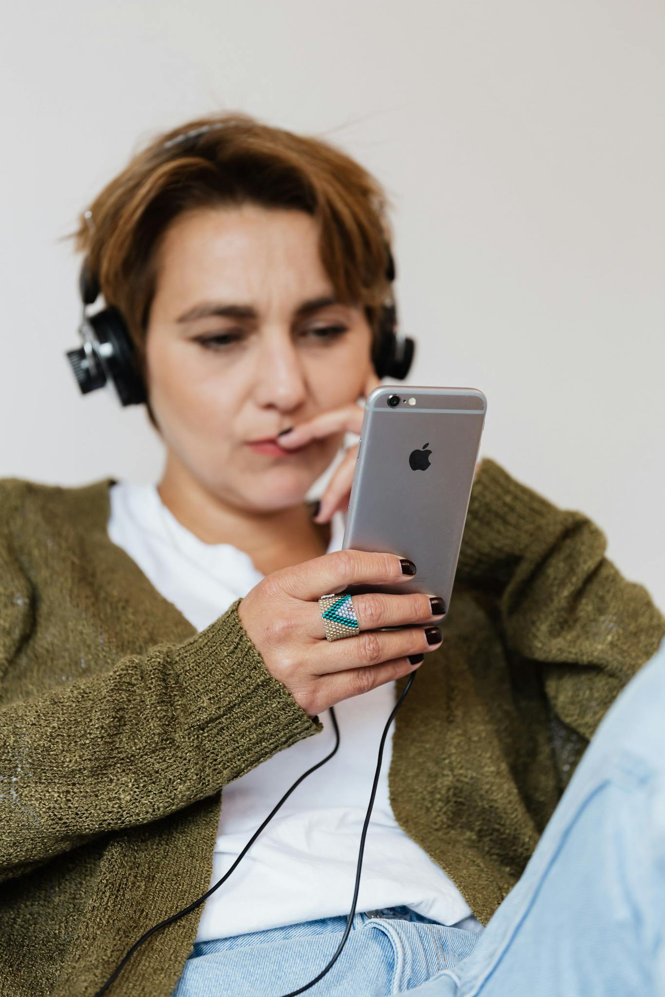 Pensive female in casual warm clothing sitting against white wall and touching face while browsing modern smartphone and listening to good music in wired headphones